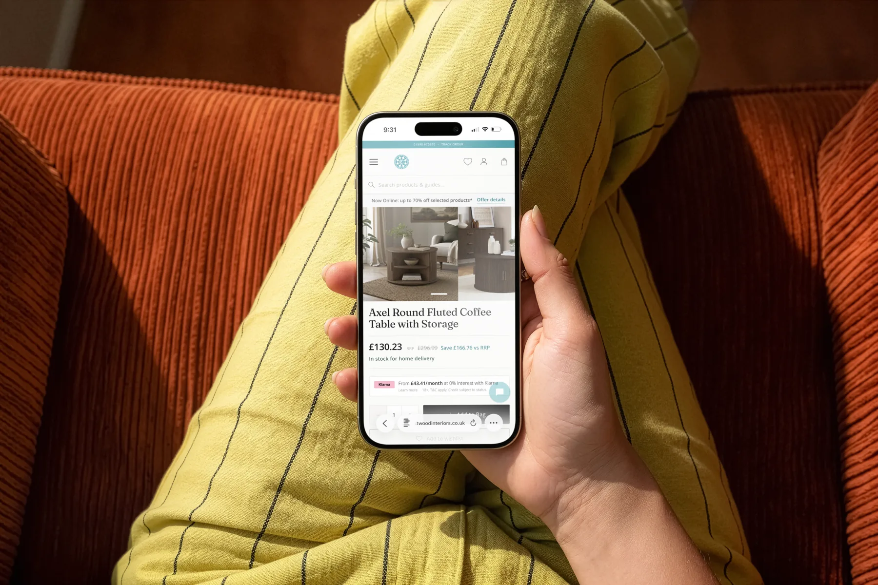Hand holding a phone over mustard pinstripe trousers and a rust corduroy sofa, showing the Crestwood Interiors product page for the Axel Round Fluted Coffee Table