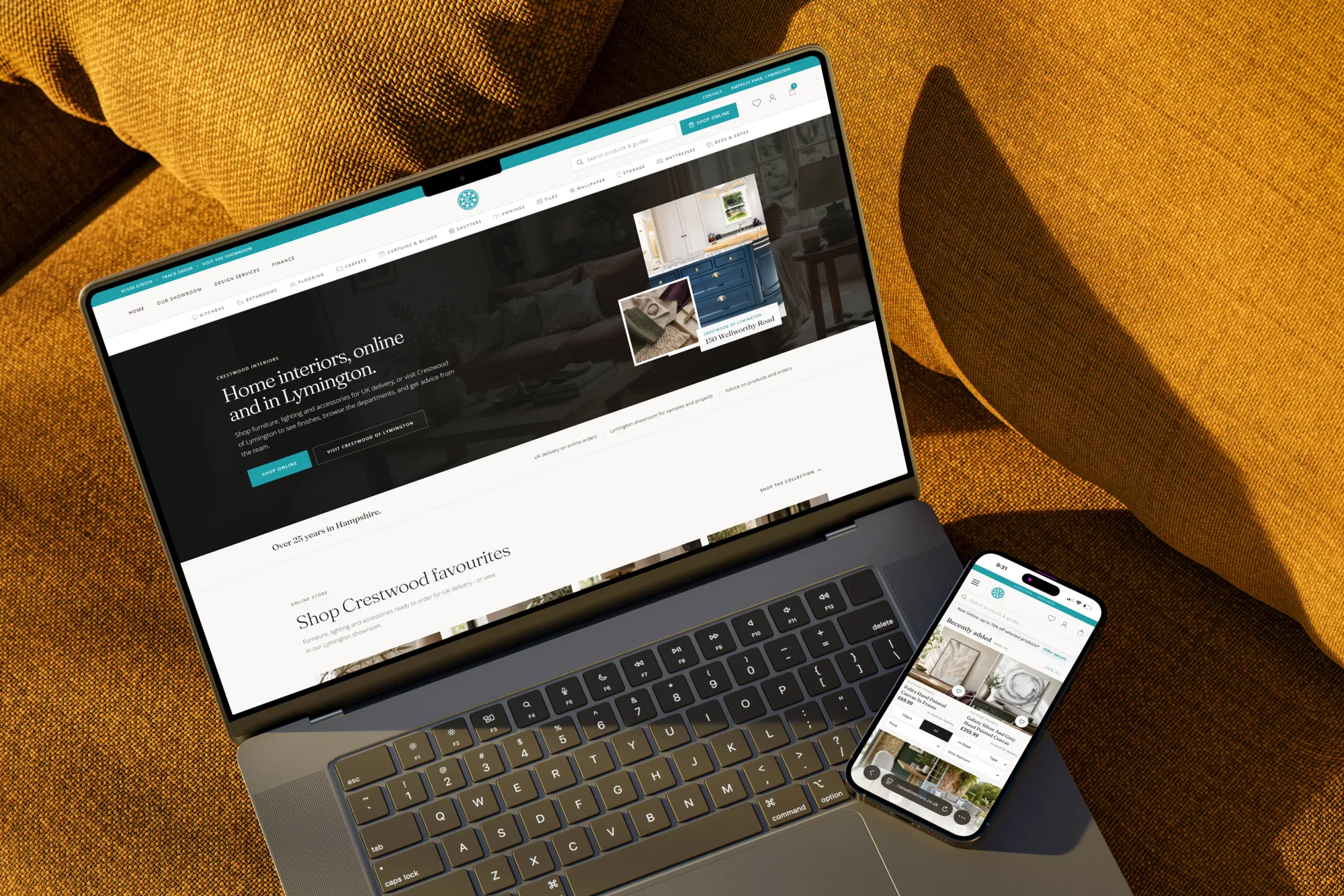 Laptop and phone on a mustard fabric sofa showing the Crestwood Interiors homepage: full-bleed hero, centred teal logomark, and equal Shop Online and Visit Crestwood of Lymington actions
